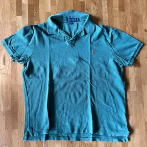 Turkos pikétröja från Ralph Lauren XL - Snygg turkos pikétröja från Polo Ralph Lauren i storlek XL, slim fit. Klassisk krage, korta ärmar och den ikoniska broderade loggan i orange på bröstet. Tillverkad i bomull som känns skön mot huden. Perfekt för en fräsch och stilren look.