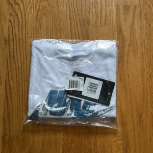 Vit Nike t-shirt med blå logga - Snygg vit t-shirt från Nike med stort blått och vitt Nike-tryck framtill. Klassisk rund hals och korta ärmar. Perfekt för en sportig och avslappnad stil. Tillverkad i mjuk bomull för skön känsla hela dagen.