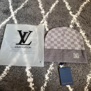 L-mössa  - Snygg grå mössa från L med klassiskt rutigt mönster och L-logga framtill. Mössan har uppvikt kant och är tillverkad i mjukt stickat material som känns lyxigt och bekvämt. Perfekt accessoar för att lyfta din vinterstil.