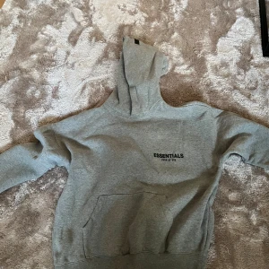 Grå Essentials Fear of God hoodie - Säljer en grå hoodie från Essentials Fear of God med svart logga på bröstet och ryggen. Storlek S, nästan helt ny andledning vrf den säljs är eftersom den inte passar, skriv gärna vid funderingar