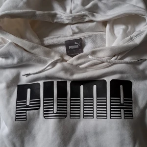 Vit hoodie från Puma med stor logga - Säljer en vit hoodie från Puma i storlek L. Tröjan har en stor svart Puma-logga framtill, huva med snörning och en klassisk känguruficka. Perfekt för dig som gillar sportig och avslappnad stil.
