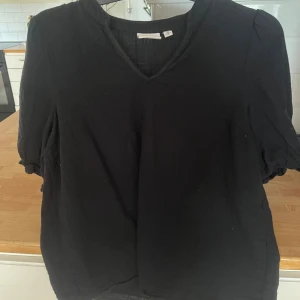 Svart blus med v-ringning från Vila - Svart blus från Vila i storlek 34. Blusen har en snygg v-ringning och korta ärmar med lätt puff och resår vid ärmsluten.