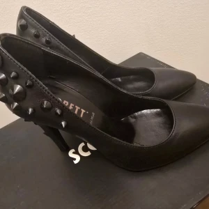Svarta pumps med nitar från Scorett - Svarta pumps i skinn från Scorett med spetsig tå och hög klack. Baksidan och klacken är dekorerade med svarta nitar för en edgy look. Perfekta för dig som vill sticka ut med en cool detalj. Klassisk form med modern twist.