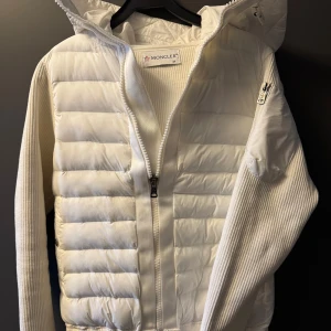 Moncler cardigan  - Säljer en snygg vit cardigan från Moncler i storlek M (helt ny). Jackan har stickade ärmar, quiltad front, dragkedja framtill och en praktisk huva. Moncler-logga på ärmen (Nfc-scan funkar) och cool detalj med ficka på sidan. Perfekt för kyliga dagar när du vill vara både varm och stilren. Priset kan diskuteras vid snabb affär. 