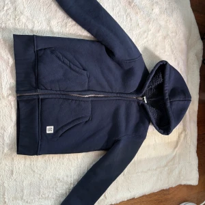 Mörkblå hoodie från Lindex med dragkedja - Mysig mörkblå hoodie från Lindex med fluffigt foder i huvan och praktisk dragkedja framtill. Tröjan har två stora fickor, ribbade muddar och lång ärm. Perfekt för kyliga dagar och enkel att matcha med jeans eller mjukisbyxor.