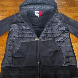 Mörkblå Tommy Hilfiger cardigan  - Mörkblå cardigan från Tommy Hilfiger med quiltad framsida och huva. Jackan har dragkedja, två fickor med knappstängning och ribbad nederkant med Hilfiger-logga. Materialmix av polyester och bomull, samt långärmad design med logotyp på ärmen. Storlek L. Nypris 2300kr mitt pris 800, kan gå ned i pris vid snabb affär!!