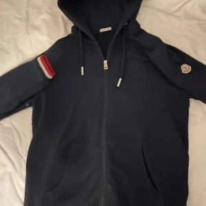 Mörkblå Moncler hoodie med dragkedja - Äkta moncler kofta som är i färgen mörkblå. Koftan kan användas i både vintern och sommaren och sitter perfekt på en. Dragkedjan funkar men du kan inte öppna den för den är sönder där nere så du sätter på dig den som en tröja men efter de kan du röra dragkedjan hur mycket du vill bara inte öppna den. Koftan är i storlek M men passar även folk med storlek S. Koftan kostar 4300 på affär. Pris kan diskuteras så tveka inte på att höra av er. 