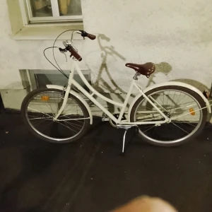 Stålhästen Damcykel - Stilren vit Stålhästen damcykel med brunt sadel och handtag. Klassisk design, fotbroms och stänkskärmar. Cykeln ser välvårdad ut med minimalt slitage, perfekt för stadskörning. Sadel och detaljer i retrostil. Inga synliga defekter.