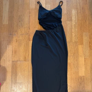 Svart långklänning med smala axelband S - Stilren svart långklänning med smala justerbara axelband och v-ringad överdel. Klänningen har en figurnära passform och är tillverkad i ett mjukt, stretchigt material. Snygg drapering över bysten och midjan ger en modern look.