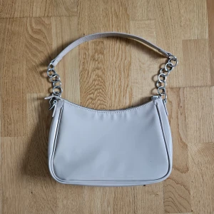 Beige axelväska från H&M Divided. - Snygg beige axelväska från H&M Divided med silvriga kedjedetaljer på axelremmen. Väskan har en stilren, minimalistisk design och stängs med dragkedja. Tillverkad i syntetmaterial och perfekt för att lyfta din outfit.