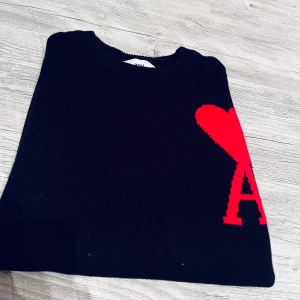 Svart stickad tröja Ami Paris hjärta - Svart stickad tröja från Ami Paris med rött hjärta och bokstaven A broderat på bröstet. Klassisk rund halsringning och långärmad modell. Perfekt statement-plagg med ikonisk design.//Tagdrop