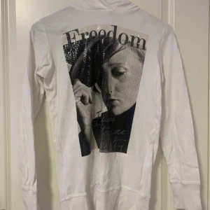 Vit hoodie med tryck och paljetter - Vit hoodie med stort svartvitt tryck och texten 'Freedom' på ryggen. Trycket har paljett-detaljer som ger en glittrig effekt. Tröjan har långa ärmar och huva 