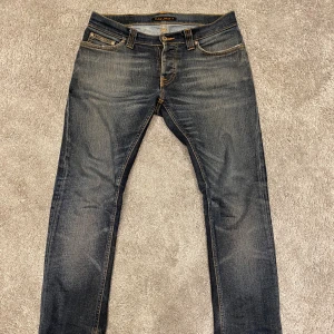 Nudie "grim tim" Jeans - Snygga mörkblå jeans från Nudie Jeans med klassisk femficksdesign. Jeansen har slitningar och kontrastsömmar, samt Nudie-logga på bakfickan. Perfekt för dig som gillar en smal siluett och cool tvätt.