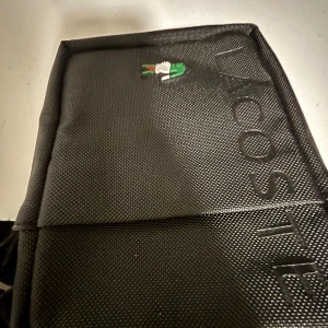 Svart axelväska från Lacoste - Snygg svart axelväska från Lacoste med klassisk krokodillogga och präglad Lacoste-text på framsidan. Väskan har en stilren, rektangulär form och är tillverkad i slitstark syntet. Perfekt för dig som vill ha en clean och sportig look.