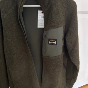 Grön teddyjacka från Lundhags (oanvändk)  - Säljer en mörkgrön teddyjacka - helt oanvänd med taggen kvar -  från Lundhags i storlek M. Jackan har en mysig pile-yta, hög krage och en praktisk bröstficka med dragkedja och patch. Perfekt för kalla dagar och outdoor-häng. Färgen heter Forest Green och materialet är ullblandning. Kvitto finns på begäran. 