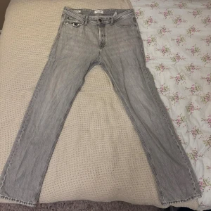 Grå jeans från Jack & Jones relaxed fit - Grå jeans från Jack & Jones med relaxed fit och klassisk femficksdesign. Jeansen har en ljusgrå tvätt och en liten sliten detalj vid myntfickan. Rak passform och normal midja, perfekta för en chill och avslappnad stil. 32W, 34L