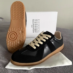 Maison Margiela skor - Maison Margiela sneakers i svart skinn och mocka med beige snörning och gummisula i naturfärg. Klassisk rund tå och platt sula, med diskret logotyp på tungan. Snygg retrostil som funkar till jeans eller dressat.