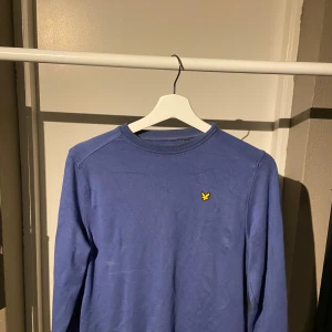 Blå långärmad tröja från Lyle & Scott - Snygg blå långärmad tröja från Lyle & Scott med klassisk gul fågel-logga på bröstet. Tröjan har rund hals och är gjord i mjuk bomull, perfekt för dig som gillar stilrena och enkla plagg. Passar dig som vill ha en clean look med en touch av färg.
