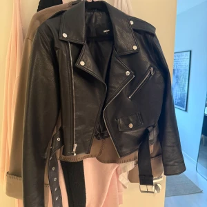 Svart skinnjacka från Bik Bok - Säljer en svart skinnjacka från Bik Bok med klassisk bikerstil. Jackan har silvriga detaljer, flera dragkedjor, knappar och ett bälte i midjan. Croppad passform och cool look som passar till många outfits. Perfekt för dig som gillar edgy stil.