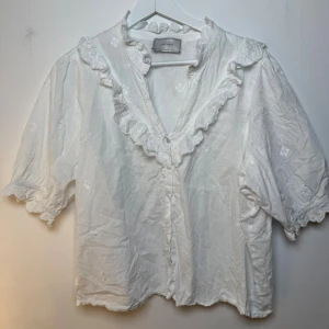 Neo Noir blus  - Vit blus med broderade detaljer och volanger runt halsringningen och ärmarna. Blusen har en v-ringning och knäpps med knappar framtill. Kort puffärm. Storlek 42/L
