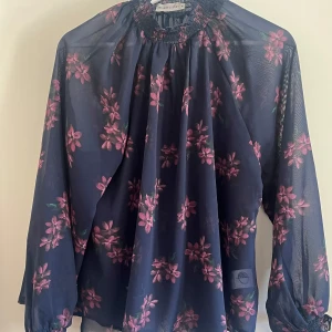 Blommig blus  - Säljer en transparent mörkblå blus med rosa blommor och gröna blad. Blusen har långärmade puffärmar, smockad halsringning och ett luftigt, loose fit. 
