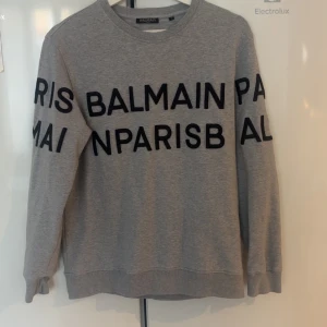 Balmain tröja (ÄKTA) - Säljer en grå sweatshirt från Balmain Paris med svart tryck över hela tröjan. Tröjan har rund halsringning, långa ärmar och är tillverkad i mjuk bomull. Perfekt för en chill och trendig look. Inga defekter mycket fint skick, tröjan säljs inte längre lyckades få tag på den genom vestaire collective 2022. Men bara legat och knappt används. Kom privat för mer info!