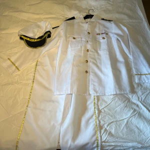 TOP GUN Kapten Vit sjöofficerskostym med guldiga detaljer - Snygg vit sjöofficerskostym med guldiga knappar, axelband och dekorativa band längs ärmar och byxor. Ingår matchande vit keps med svart och guld, samt skjorta med bröstfickor och medaljdetaljer. Perfekt för temafest eller maskerad.