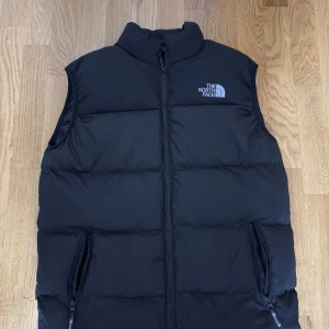 Svart dunväst från The North Face - Svart dunväst från The North Face med hög krage och två praktiska fickor med dragkedja. Västen har en quiltad design och logotypen i vitt på bröstet. Perfekt för lager-på-lager och chill stil under kyliga dagar.