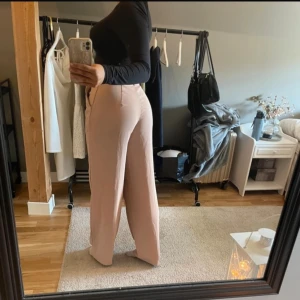 Beiga vida kostymbyxor - Säljer ett par snygga beiga kostymbyxor med hög midja och vida ben. Byxorna har klassisk knapp och dragkedja framtill samt hällor för bälte. Perfekta för en clean och stilren outfit.