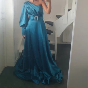 Blå långklänning med slits och bälte Aldrig använt!! - Säljer en blå långklänning i glansigt material med en axel, puffärm och bredt bälte med glittrande spänne. Klänningen har en hög slits och en elegant, svepande kjol. Perfekt för bal eller festliga tillfällen.