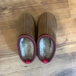UGG bruna slip-in mockatofflor - Bruna slip-in tofflor från UGG med mjuk och fluffig insida i vitt. Yttermaterialet är mocka och de har en platt sula. Runt öppningen finns ett dekorativt mönster i rött och beige samt en röd detalj baktill. Perfekta för att hålla fötterna varma och sköna.