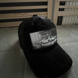 Svart meshkeps med biltryck - Svart keps med mesh på baksidan och justerbar snapback. Framtill finns ett stort svartvitt tryck med motiv av en klassisk sportbil framför en byggnad. Kepsen har böjd skärm.