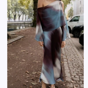 Lång omlottklänning i sidenlook från Zara - Snygg långklänning med svepande siluett i glansigt material. Klänningen har ett unikt mönster i blått, vinrött och vitt, samt en axelbandslös design med vida ärmar och omlott-detalj. Perfekt för dig som vill sticka ut med en cool och elegant stil.