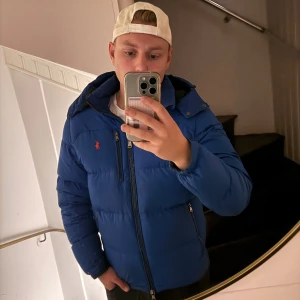 Blå pufferjacka från Polo Ralph Lauren - Blå pufferjacka från Polo Ralph Lauren med orange logga på bröstet. Jackan har huva, flera dragkedjor och quiltad design. Materialet är syntet och dun, perfekt för kalla dagar. Snygg och sportig look med lång ärm och elastiska muddar. Tecken på användning men inget som stör helheten 