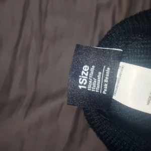 Svart Nike mössa med broderad logga - Svart ribbstickad mössa från Nike med klassisk uppvikt kant och vit broderad Nike-logga framtill. Perfekt för kalla dagar och enkel att matcha med olika outfits. Tillverkad i mjukt och stretchigt material för skön passform.