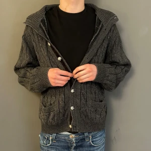 Grå stickad kofta med knappar - Snygg grå stickad kofta med ribbade muddar och framfickor. Koftan har ett randigt mönster och stora silvriga knappar framtill. Den har en bred krage och långärmade ärmar, perfekt för lager på lager under höst och vinter.