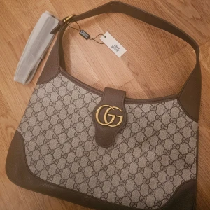 Axelväska med GG-mönster - Snygg axelväska i beige och brunt med klassiskt GG-mönster och guldfärgad logga framtill. Väskan har bred axelrem, detaljer i skinn och praktiska innerfack med dragkedja. Perfekt för dig som vill ha en ikonisk och trendig accessoar.