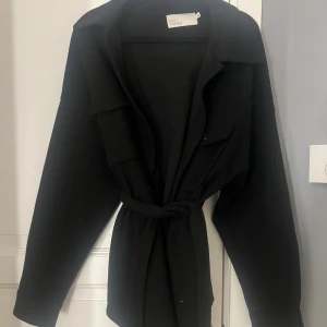 Svart overshirt från NLY Trend - Svart overshirt med klassisk krage och stora bröstfickor, knäppning framtill med svarta knappar och tillhörande bälte i midjan. Jackan har lång ärm och en loose passform, perfekt för lager-på-lager. Materialet är mjukt och känns som ull.