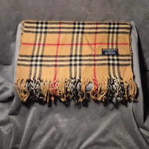 Rutig beige halsduk från Burberry - Klassisk halsduk från Burberry i beige med rutigt mönster i svart, vitt och rött. Gjord av 100% lammull och har fransar i ändarna. Perfekt accessoar för att lyfta din outfit med en tidlös brittisk vibe.