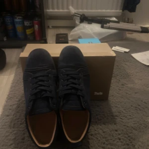 Christian Louboutin mörkblå sneakers mocka - Säljer ett par mörkblå sneakers från Christian Louboutin i lyxig mocka med klassisk röd sula. Skorna har snörning, rund tå och diskret broderad logga på hälen. Insidan är beige och sulan är robust. Perfekta för dig som vill sticka ut med exklusiv stil. Erbjuder tvättning vid snabbt köp