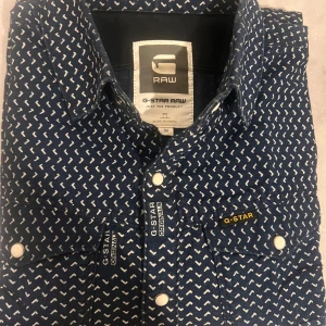Blå mönstrad skjorta från G-Star RAW - Säljer en blå skjorta från G-Star RAW med vitt grafiskt mönster, klassisk krage och vita tryckknappar. Skjortan har två bröstfickor med lock och märkets logga på ena fickan. Tillverkad i bomull och har lång ärm.
