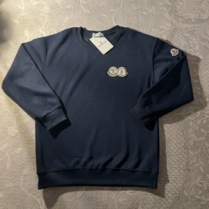 Mörkblå sweatshirt från Moncler - Snygg mörkblå sweatshirt från Moncler med klassisk rund halsringning och broderad logga på bröstet samt ärmen. Tröjan har ribbade muddar och är tillverkad i mjuk bomull, perfekt för en clean och stilren look.