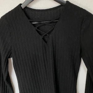 Svart ribbad topp med snörning från SHEIN - Svart långärmad topp från SHEIN i ribbad polyester med snygg snörningsdetalj vid v-ringningen. Modellen är figurnära och har en enkel, stilren look som passar till många outfits.