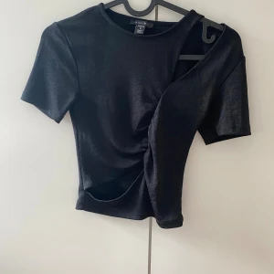 Svart topp från AMIS med twistdetalj - Säljer en svart topp från AMIS i storlek S. Toppen har korta ärmar, rund halsringning och en snygg twistad detalj framtill som ger en cool look. Materialet är stretchigt och följsamt, perfekt till jeans eller kjol.