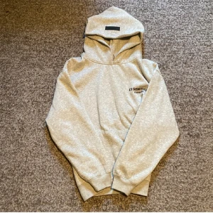 Grå Essentials Fear of God hoodie - Snygg grå hoodie från Essentials Fear of God med svart logotyptryck både fram och bak. Tröjan har huva, långa ärmar och ribbade muddar. Perfekt för dig som gillar streetwear och vill ha en clean look. Materialet är mjuk bomullsmix som känns skön mot huden.