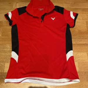 Victor - Röd pikétröja - Badminton  - Sportig röd pikétröja från Victor med svarta och vita detaljer på sidorna och ärmarna. Tröjan har korta ärmar, krage och knappar framtill. Perfekt för dig som gillar att sticka ut på träningen eller på tennisbanan.