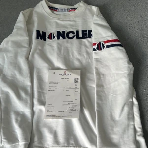 Vit långärmad tröja från Moncler - Vit långärmad tröja från Moncler med broderad logga på bröstet och coolt band med logga och röd/blå detalj på ena ärmen. Tröjan har rund hals och är gjord i mjuk bomull. Perfekt för dig som gillar stilrena märkesplagg med snygga detaljer.