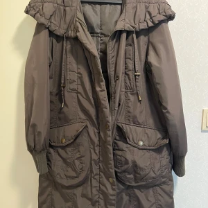 Brun parkajacka från XINT med huva - Parkajacka i brun nyans från XINT med stor huva, snörning och dragkedja framtill. Jackan har stora fickor med lock och tryckknappar, ribbade muddar vid ärmslut och justerbar midja baktill. Perfekt för kyliga dagar och har en avslappnad passform.