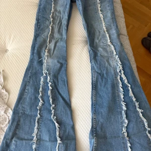 Blå bootcut jeans med fransade detaljer - Säljer ett par ljusblå jeans med bootcut passform och fransade sömmar längs benen. Perfekt för dig som gillar en unik stil. Vid funderingar hör av er💕