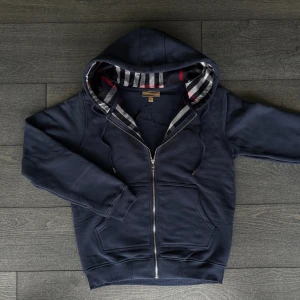 Marinblå hoodie från Burberry - Säljer en marinblå hoodie från Burberry med dragkedja och klassiskt rutigt foder i huvan. Tröjan har snörning vid huvan, två fickor fram och ribbade muddar. Perfekt för dig som vill ha en stilren och lyxig look.